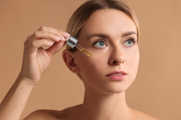 Beautiful woman applying cosmetic serum onto her face on beige background, closeup