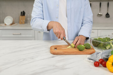 Man cutting fresh broccoli with knife near containers at white marble table in kitchen, closeup and space for text. Food storage