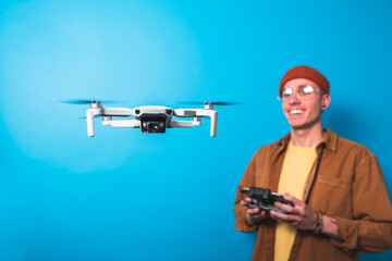 Smiling young man piloting a drone in flight with a remote control on an isolated blue background. Drone pilot and aerial filming concepts. Focus on the drone