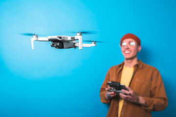 Smiling young man piloting a drone in flight with a remote control on an isolated blue background. Drone pilot and aerial filming concepts. Focus on the drone