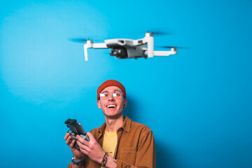 Smiling young man learning piloting a drone in flight with a remote control on an isolated blue background. Drone pilot and aerial filming concepts. Focus on the drone