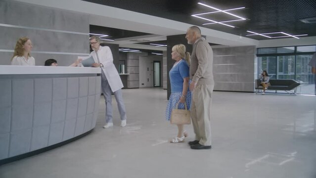 Elderly couple come to reception desk in clinic lobby entrance and make appointment with doctor. Female administrator talks with patients at information counter. Medical staff work in modern hospital.