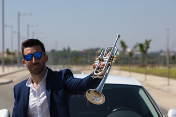 handsome young man with beard, blue suit and sunglasses on his white car shows his trumpet in a happy attitude. The man is a musician. October 1st international day of music. © @skuder_photographer