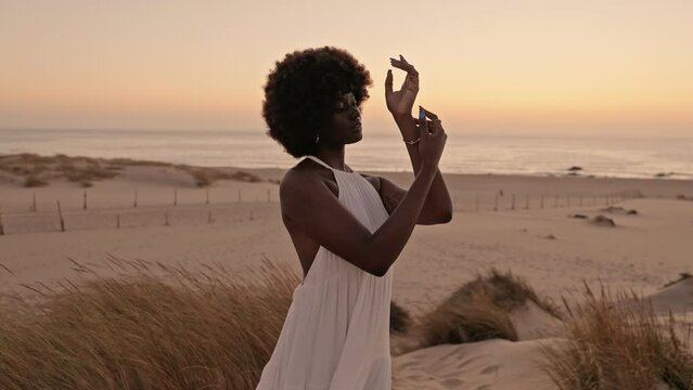 A beautiful black woman in a flowing white dress performs a conceptual dance on sandy beach dunes at sunset, connecting harmoniously with nature's rhythm and beauty