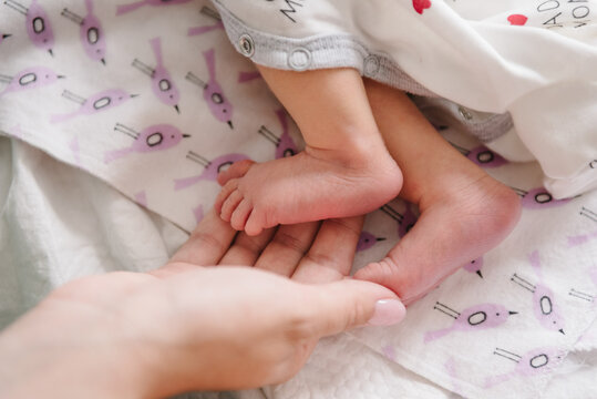 Baby feet. Young mother holding child's feet. Cute little baby lying on bed at home. Mom hold son legs. Closeup. Tiny, bare feet of newborn girl or boy wrapped in hands. Sleeping daughter. Top view
