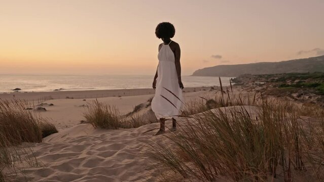 Wanderlust as a beautiful black woman with an afro hairstyle strolls barefoot along the beach at sunset, embracing the tranquility of soft sand wearing an airy white dress.
