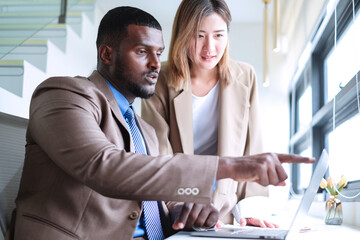 Black businessman pointing at laptop screen to asian businesswoman colleague looking at graphs and company sales data.