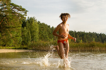 A slender guy child teenager runs along the water along the coast at sunset, splashing from the water.