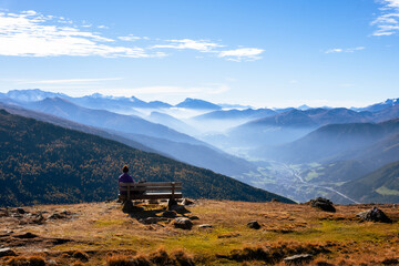 Naklejka premium A woman traveler is sitting on a bench with a panoramic mountain view on the trail in Austrian Alps
