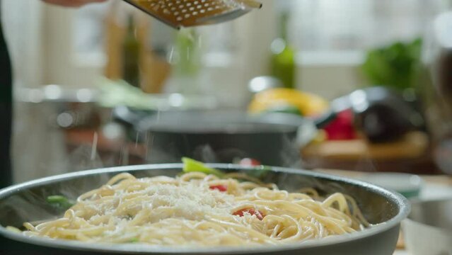 Close-up impersonal shot of grating cheese over hot spaghetti with veggies in frying pan while serving just cooked pasta dish