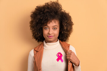 confident black young woman showing pink ribbon in beige background. breast cancer, awareness concept.