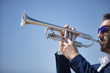 Young Hispanic man, wearing a jacket and sunglasses, playing a pretty, silvery trumpet outdoors....