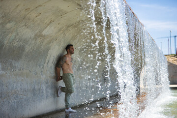 Young, attractive and Hispanic man, shirtless and with a muscular body, leaning against a wall under a bridge where a waterfall falls. Concept, beauty, water, masculinity.