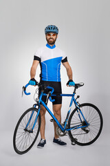 Happy male cyclist in cycle kit standing with bike in light studio. Front view of bearded bicyclist in blue jersey smiling to camera, isolated on grey background. Concept of cycling sport, activity.
