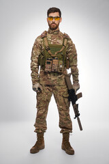 Serious male soldier in body armor holding rifle, while posing indoors. Front view of dark-haired...
