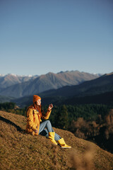 Woman full-length sitting resting on a hill and looking at the mountains in a yellow raincoat and jeans happy camping trip in the autumn, freedom lifestyle 