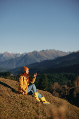 Fototapeta premium Woman full-length sitting resting on a hill and looking at the mountains in a yellow raincoat and jeans happy camping trip in the autumn, freedom lifestyle 