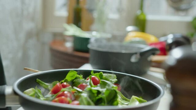 Hands of male chef cooking fresh spinach and tomatoes in a frying pan, using wooden spatula to stir vegetables and adding salt. Close-up shot