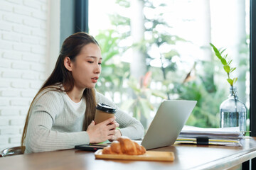 Asian woman with tired face editing work on vacation to relax There is coffee on the table, drinks, snacks, documents, a hand on a coffee cup symbolizes boredom.