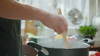 Unrecognizable man in apron stirring boiling food in cooking pot with wooden spatula in the kitchen. Close-up shot, side view