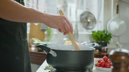 Male chef in apron stirring boiling food in a pot with wooden spatula while preparing dinner in the kitchen. Cropped shot, side view - Powered by Adobe