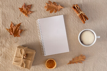 Fototapeta na wymiar Autumn composition with an empty blank, a cup of coffee, a gift wrapped in craft paper, dry leaves, dried orange, cinnamon on a background of linen fabric. Flat lay, copy space, fall concept.