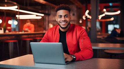 Fototapeta premium young arab man in a red pullover jacket, freelancer or student working on a laptop in a cafe at the table. Black IT professional working remotely using laptop while sitting in cafe