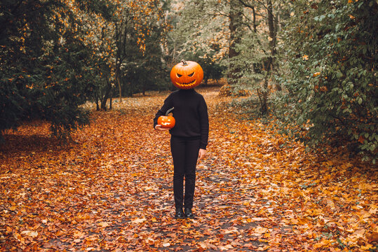 Halloween. Pumpkinhead. A man with a pumpkin on his head stands in a park on an alley strewn with yellow autumn leaves.