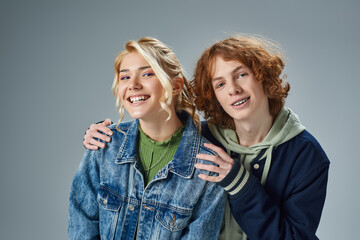 redhead teenage boy embracing shoulders of cheerful blonde girlfriend on grey, youth fashion