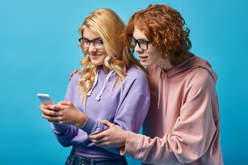 joyful blonde teen girl in eyeglasses and trendy hoodie using smartphone near redhead friend on blue