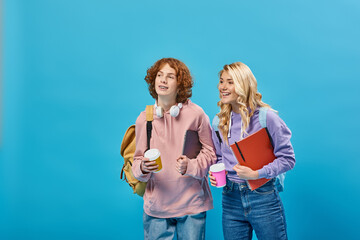 Fototapeta na wymiar joyful teenage students with backpacks and takeaway drinks in paper cups looking away on blue