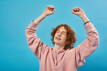 smiling and dreamy redhead teen guy in hoodie standing with closed eyes and raised hands on blue