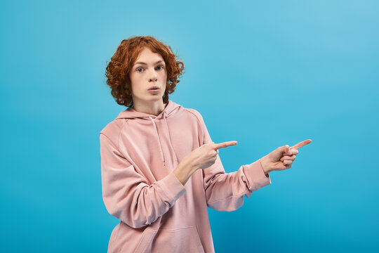 surprised redhead teen boy in trendy hoodie looking at camera and pointing away with fingers on blue