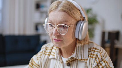 Senior woman wearing headphones looks at computer screen in her home office, freelancer's lifestyle