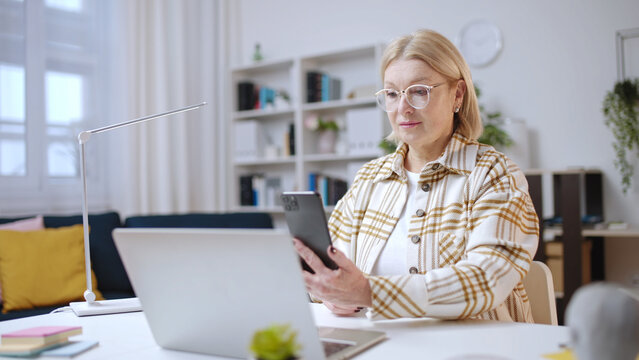 Woman in her 50s using smartphone to make a purchase, scanning qr code on laptop