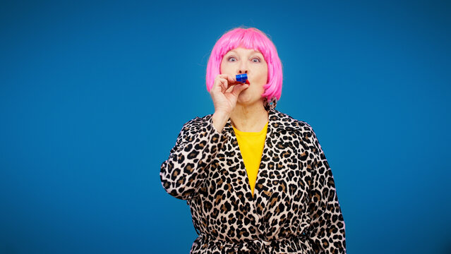 A fashionable senior woman in a pink wig is blowing a party horn, celebrating a holiday