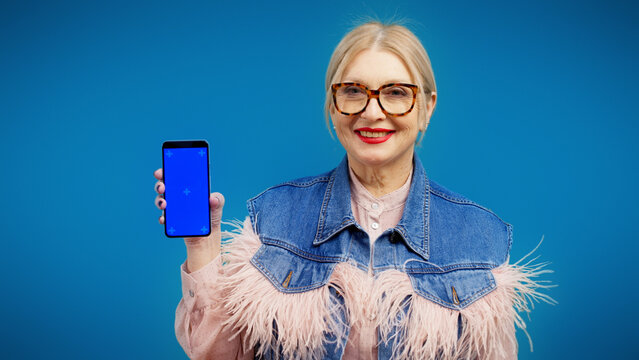 A smiling senior woman in trendy clothes is showing a smartphone with a pre-keyed screen