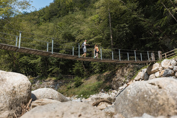 Fototapeta na wymiar Young heterosexual hiking couple crossing the suspension bridge over a mountain river during summer hiking tour