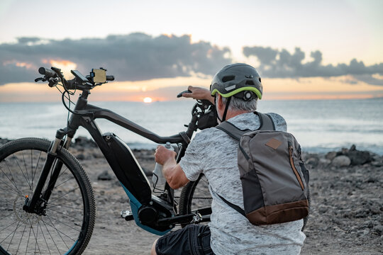 Rear view of active caucasian senior man with electric bicycle at sunset on the beach - elderly man with helmet enjoying healthy lifestyle