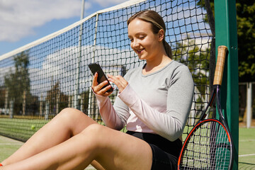 Young smiling female tennis player sitting on court and using smart phone during a break in training