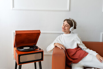 Beautiful teenager boy wearing earphones smiling and listening music near wooden vinyl record player. Minimalist home interior, retro aesthetic