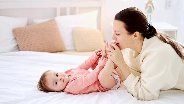 mom kisses the legs of a small child with her daughter, play on the bed at home, mom and baby have fun together, happy motherhood