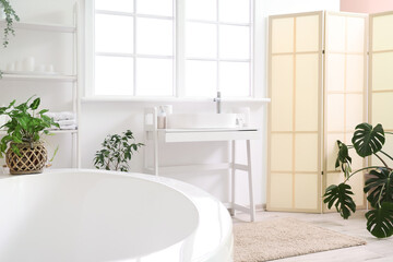 Interior of light bathroom with sink, bathtub, shelving unit and houseplants