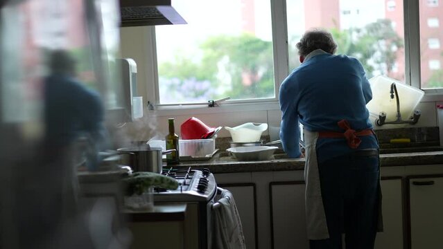 Senior By Kitchen Sink Washing Dishes And Preparing Food At Stove. Elderly Person Cooking Food Wearing Apron, Candid Genuine Retired Lifestyle