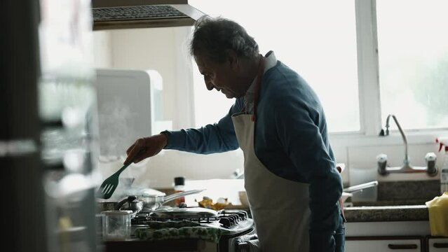 Senior chef cooking food by kitchen stove stirring meal inside pan, Older Man's Detailed food preparation Moment Near sink