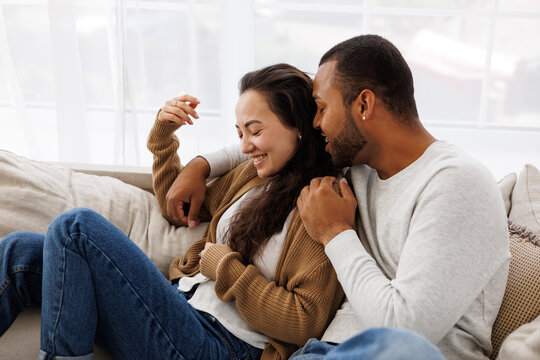 Smiling african american man hugging and talking to asian girlfriend while relaxing on couch at home