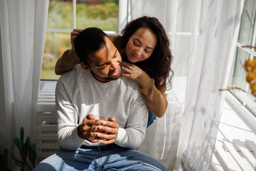 Smiling asian woman hugging african american boyfriend with coffee near window and sunlight at home