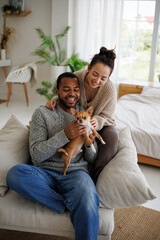 Happy young african american man in sweater holding chihuahua dog near asian girlfriend on armchair at home
