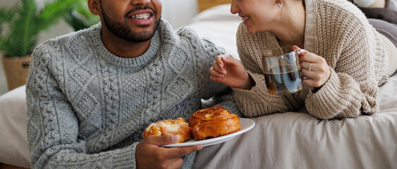 Banner image of smiling multiethnic couple in sweaters holding coffee and sweet pastry in bedroom