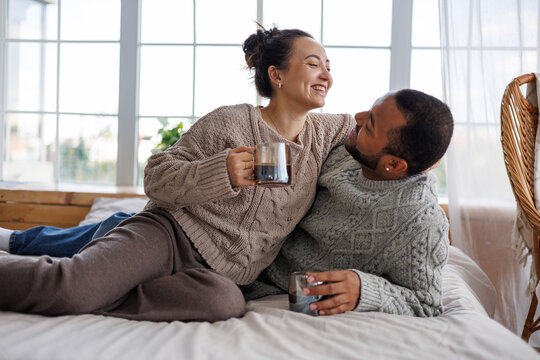 Cheerful young multiethnic couple in sweaters holding coffee while hugging and relaxing on bed at home